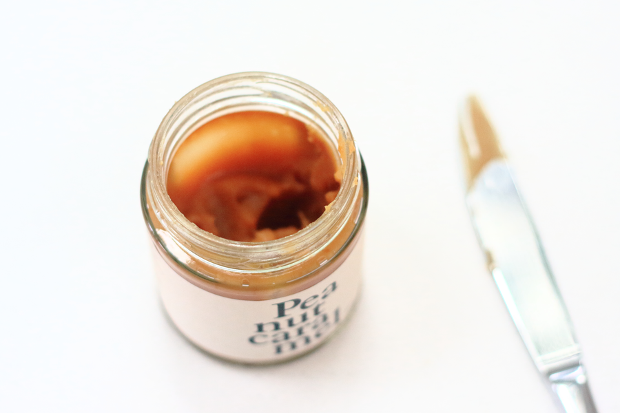 A photo of a jar of peanut caramel on a white surface, taken from an angle in natural light, with a knife alongside